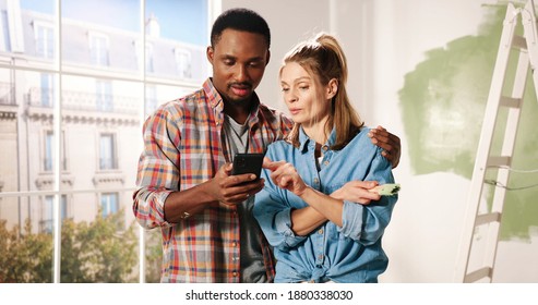 Joyful Multi-ethnic Young Married Couple Caucasian Wife And African American Husband Hugging Standing In Room Typing On Mobile Phone Choosing Design Of New Home. House Repair. Renovation Concept