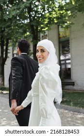 joyful muslim bride in wedding dress holding hands with groom in suit