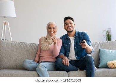 Joyful muslim couple watching funny TV programs, sitting on couch at home, eating popcorn. Laughing arab man holding remote and happy woman in hijab pointing at TV screen, copy space