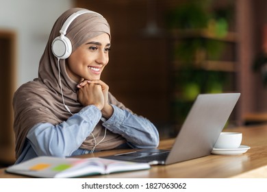 Joyful Muslim Woman In Hijab Studying Online At Cafe, Using Laptop And Headset, Free Space. Happy Female Student Having Online Lesson, Looking At Laptop Screen And Smiling, Online Education Concept