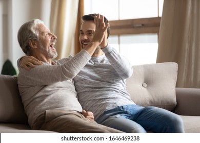 Joyful Older Mature Man Giving High Five To Happy Grownup Son, Celebrating Making Good Decision. Smiling Two Male Generations Family Feeling Excited About Good News, Resting On Sofa At Home.