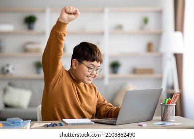 Joyful overweight chinese boy teenager using laptop at home, sitting at table among books and notebooks, looking at computer screen and raising hand up, wearing glasses, copy space