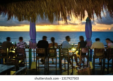 Joyful people are relaxing by the wooden rack on the terrace of the outdoor cafe on the breathtaking background of the sunset sky and the sea on Bali. Horizontal.