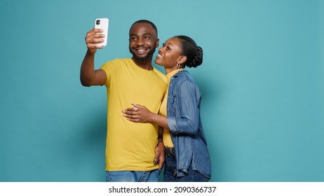 Joyful People Using Mobile Phone To Take Selfies Together In Studio. Man And Woman Taking Picture With Smartphone And Having Fun In Front Of Camera. Carefree Couple Feeling Happy.