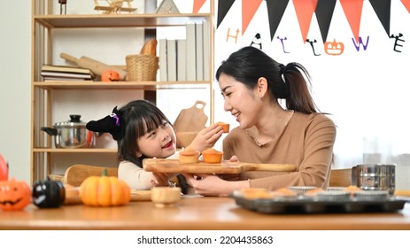 Joyful And Playful Little Asian Girl In A Halloween Costume Is Having A Great Time With Her Mom, Feeding Cupcake To Her Mom. Happy Family Time Concept