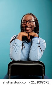 Joyful Pleased Young Adult Person With Confident Smile And Casual Looking. Excited Cheerful African American Pretty Attractive Woman Sitting On Chair While Smiling At Camera On Blue Background.