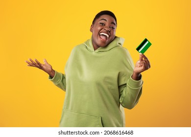 Joyful Plus Size Black Woman Holding Credit Card Smiling To Camera Posing Over Yellow Studio Background. Excited Customer Showing Her Bank Card Advertising Payment Service Concept