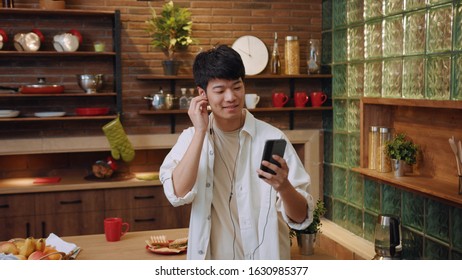 Joyful positive young chinese man in headphones listens to music, enters the modern high-tech kitchen, takes mug and pours out hot water in it. Making tea, enjoying lifetime, fun. Positive mood, fun