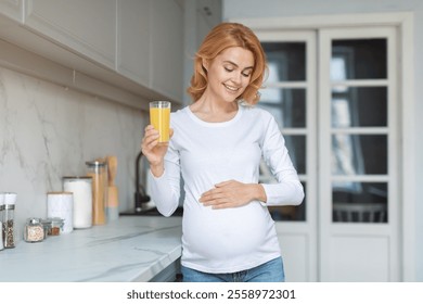 A joyful pregnant woman stands in her stylish kitchen, holding a glass of orange juice with one hand while gently resting the other on her baby bump, radiating happiness and health. - Powered by Shutterstock