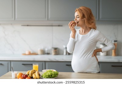 A joyful pregnant woman tastes a fresh apple while standing in a stylish kitchen. Colorful fruits and a glass of juice are laid out on the counter, promoting healthy eating habits, copy space - Powered by Shutterstock