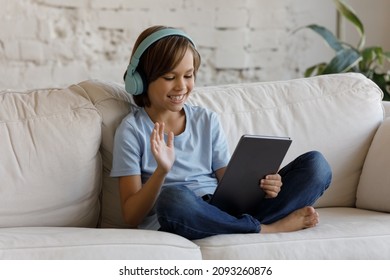 Joyful Preteen Kid Boy Wearing Wireless Headphones, Waving Hand Starting Distant Video Call Conversation Using Digital Computer Tablet, Sitting On Couch At Home, Communicating Or Studying Remotely.