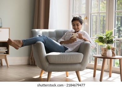 Alegre y bonita joven milenaria usuaria india de celulares descansando en un sillón en el acogedor hogar, tocando la pantalla del teléfono móvil, escribiendo, charlando en línea, disfrutando del ocio, comprando en Internet, comunicando