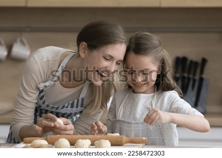 Joyful pretty mother and happy little daughter kid in aprons baking together, shaping, kneading dough, preparing pies, cookies, smiling, laughing, enjoying culinary home activity