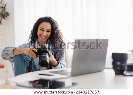 Joyful Professional Photographer Lady Holding Camera Using Laptop Sitting At Workplace Indoors. Professional Photography, Modern Arts And Creative Career Concept. Selective Focus