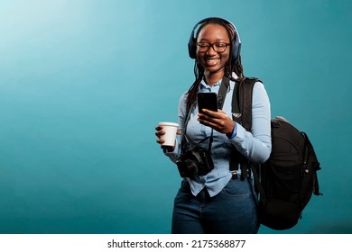 Joyful Professional Photographer With Wireless Headphones And Modern Smartphone Listening To Music On Blue Background. Happy Woman With Camera Device Enjoying Songs While Browsing Internet On Phone.