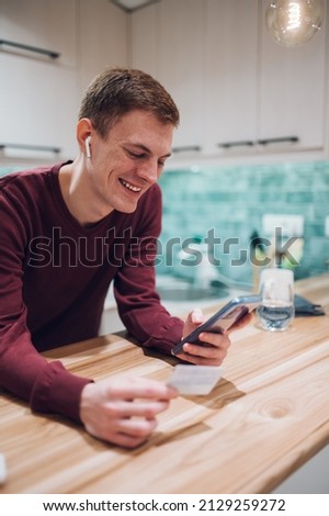 Joyful redhead positive man using a smartphone and smiling while looking at his credit card in the kitchen at home. Electronic payment. Online banking. Online shopping.
