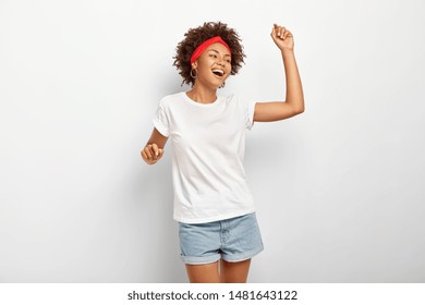 Joyful Relaxed African American Woman Moves With Rhythm Of Favourite Music, Has Happy Look, Smiles Carefree, Keeps Arms Raised, Wears Everyday T Shirt And Jean Shorts, Turnes Right, Enjoys Loud Vibes