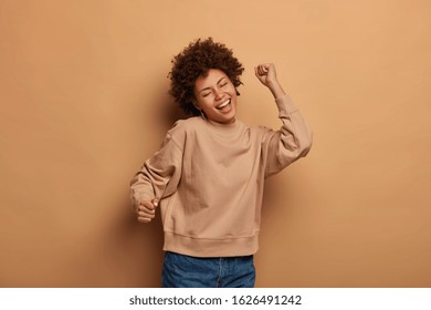 Joyful Relaxed Woman Dances Carefree Against Brown Background, Moves With Rhythm Of Favourite Music, Feels Pleased And Delighted, Wears Brown Jumper And Jeans, Being Like Winner, Enjoys Life