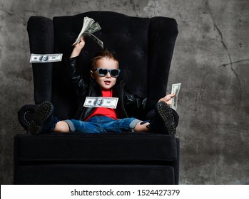 Joyful Rich Kid Boy In Sunglasses, Leather Jacket, Red T-shirt And Blue Jeans Is Sitting In Big Luxury Armchair With A Bundle Of Dollars Cash, Throwing Money, Spending