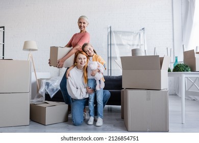 Joyful Same Sex Couple With Adopted Daughter Looking At Camera Near Carton Packages In New Home