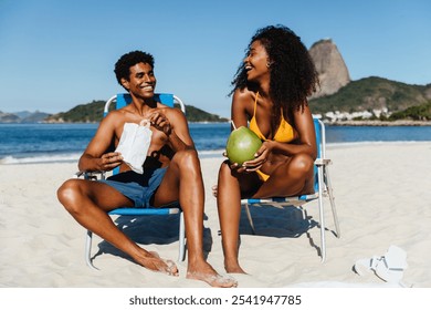 A joyful scene of two friends sitting on a sandy beach in Brazil, enjoying a sunny day. They are laughing together while having refreshing coconuts and snacks, with a scenic backdrop of mountains. - Powered by Shutterstock