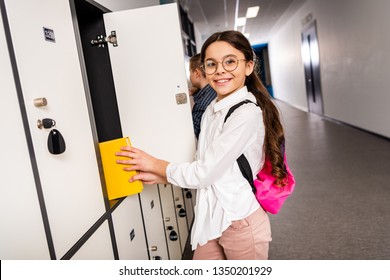 Joyful schoolgirl in glasses putting book in locker during brake in school - Powered by Shutterstock