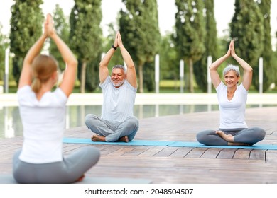 Joyful Senior Family Couple Staying In Lotus Position With Folded Hands Raised Up Of Female Yoga Trainer, Relaxed Elderly Husband And Wife Enjoying Meditation Workout Outside In City Park. Meditation 