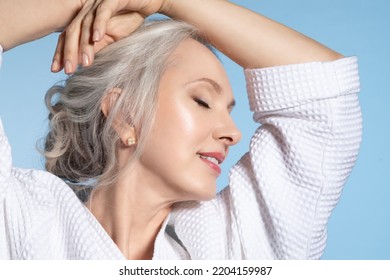 Joyful senior female in bathrobe enjoying perfect skin, close up studio portrait on blue. Smiling mature gray-haired lady closed eyes. Aged cosmetology, skincare, spa and health of elderly woman. - Shutterstock ID 2204159987