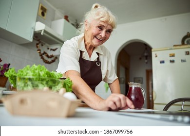 Joyful Senior Lady In Apron Searching For Recipe On The Internet And Smiling While Cooking Dinner At Home