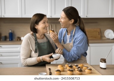 Joyful senior mom and adult daughter having fun while cooking dessert, decorating chocolate muffins, tasting cupcakes. Happy young woman giving sweet cupcake to mother for biting - Powered by Shutterstock