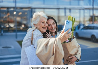 A joyful senior mother and her adult daughter pose for a selfie, phone in hand, outside a grocery store, bags of shopping in tow, showcasing intergenerational bonding. - Powered by Shutterstock