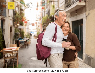 Joyful Senior Tourists Couple Hugging Posing With Backpack Holding Paper Coffee Cups On Cozy Lisbon Street With Outdoor Cafes. Retired Elderly Spouses Enjoying Vacation And Retirement In Europe