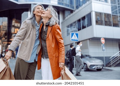 Joyful Senior Woman With Paper Shopping Bags Hug Asian Lady On Modern City Street On Autumn Day. Friends Spend Time Together