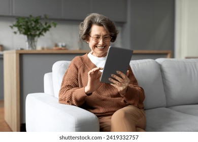 Joyful senior woman, smiling brightly and using digital tablet while sitting on cozy sofa at home, creating warm and modern lifestyle scene - Powered by Shutterstock