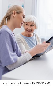 Joyful Senior Woman Smiling Near Young Nurse Holding Photo Album