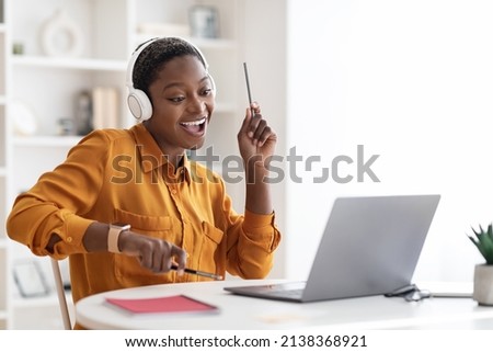 Joyful short-haired millennial black lady having break while working at office, watching video on laptop, using wireless headset, holding pens and playing imaginary drums, relaxing, copy space