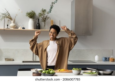 Joyful Sincere Laughing Millennial African American Woman In Eyewear Making Moves, Dancing To Entertaining Disco Music In Modern Kitchen, Distracted From Preparing Healthy Fresh Organic Salad.