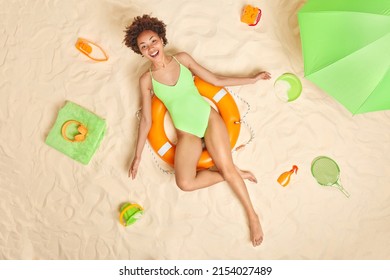 Joyful Slim Female Tourist Spends Summer Vacation At Beach Dressed In Green Swimsuit Poses On Inflated Lifebuoy Gets Suntan Surrounded By Sand Toys Green Parasol To Hide From Sun. Overhead Shot