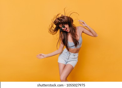 Joyful Slim Girl In Blue Shorts Dancing With Pleasure. Long-haired Young Woman In Summer Outfit Having Fun On Yellow Background.