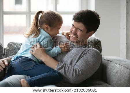Joyful small preschool baby girl tickling laughing young father, having fun together on sofa at home. Happy millennial daddy playing with overjoyed little daughter, enjoying weekend time indoors.