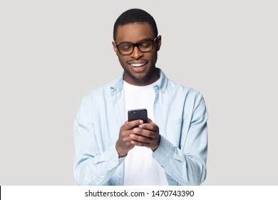 Joyful smiling african american young man in eyeglasses holding cellphone in hands, typing message, chatting with friends in social networks, playing online game, isolated on grey studio background.