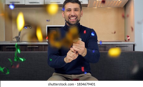 Joyful smiling bearded man celebrates victory favorite team explodes confetti cannon. Male in blue sweater sits at home on couch on background kitchen watching sports football TV
