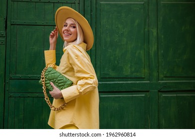 Joyful smiling woman wearing yellow hat, classic shirt, holding quilted faux leather green bag with chain, posing on green background in street. Copy, empty space for text - Shutterstock ID 1962960814