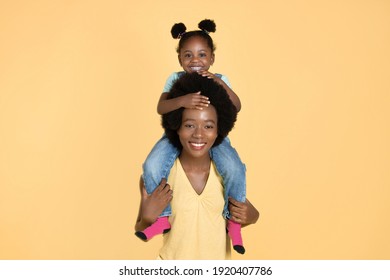 Joyful Smiling Young Black Woman Mother Giving Her Adorable Pretty Daughter Piggy Back Ride, Isolated On Yellow With Copy Space