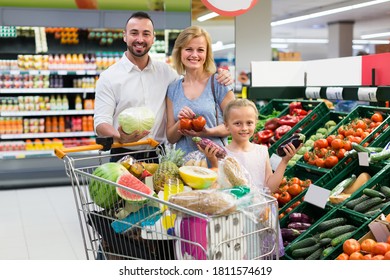 Joyful smiling young parents with little girl picking vegetables in supermarket