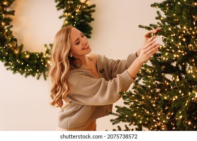 A joyful smiling young woman is preparing for the Christmas holiday, decorating a Christmas tree and holding gift boxes in a cozy interior room of the house. Selective focus