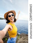 Joyful and smiling young woman traveler taking selfie with phone during summer vacation in seaside location. Vertical.