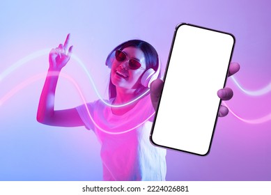 Joyful stylish young asian woman showing cell phone with white blank screen, using wireless stereo headphones, listening to music and dancing in neon light, recommending nice mobile app, collage