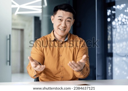 Joyful successful businessman watching in web camera, smiling man talking with colleagues remotely using video call on laptop, asian man at workplace inside office.