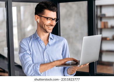 Joyful successful caucasian businessman, manager or ceo, wearing glasses, standing in office, holding and uses laptop, chatting online with colleagues or client, browsing internet, smiling - Shutterstock ID 2311823777
